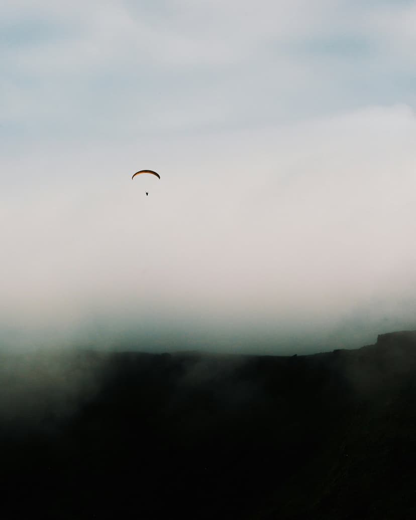 Paraglider silhouetted against a glowing sunrise sky over misty mountain peaks in a vertical aerial view.