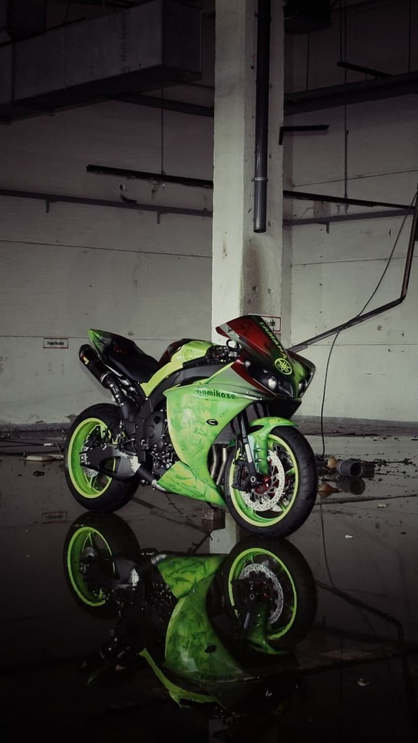 Green Yamaha sportbike with a red windshield reflecting in a puddle on a dark, gritty industrial warehouse floor.