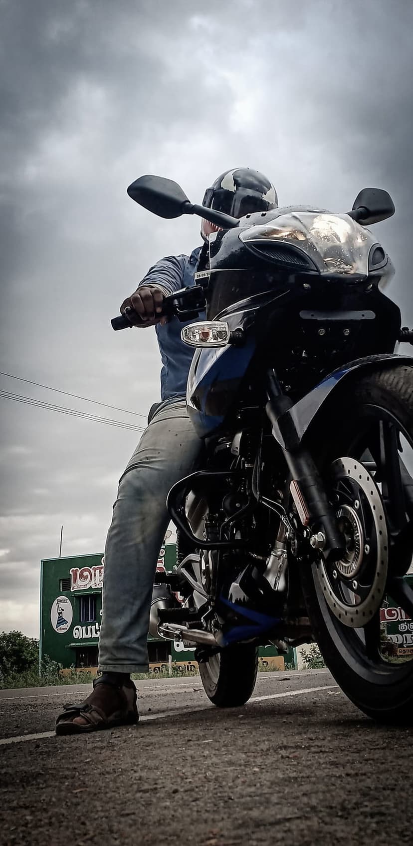 Man in a helmet rides a black motorcycle past a green building with Tamil text under a vast cloudy sky on a road.