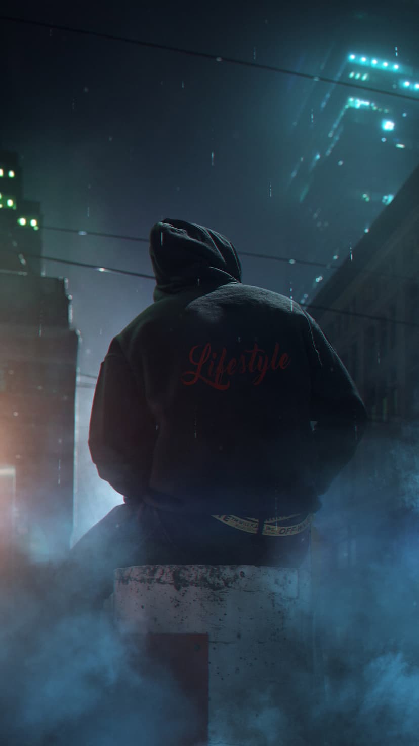 Person in a black hoodie sits on a concrete block during a rainy night with neon city lights in the distance.