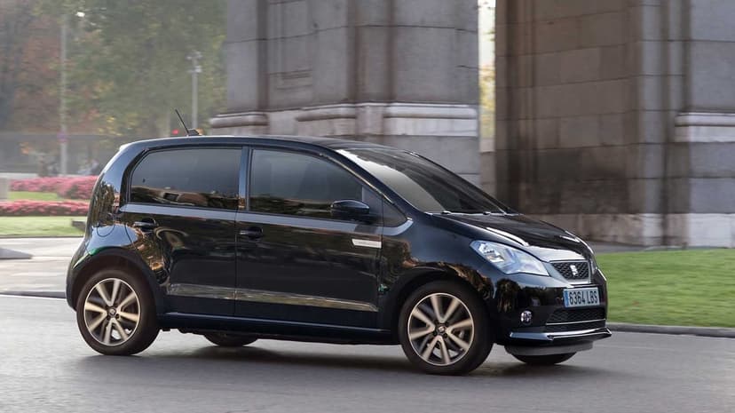 Black SEAT Mii hatchback driving past a stone archway with motion-blurred wheels on a clean city street.
