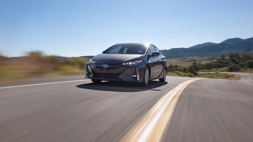 Toyota Prius Prime driving on a curved highway with motion blur against a clear blue sky and rolling hills.