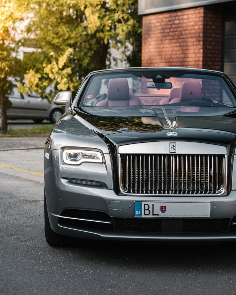 Silver Rolls-Royce convertible featuring a red leather interior and chrome grille parked under sunlit green trees.