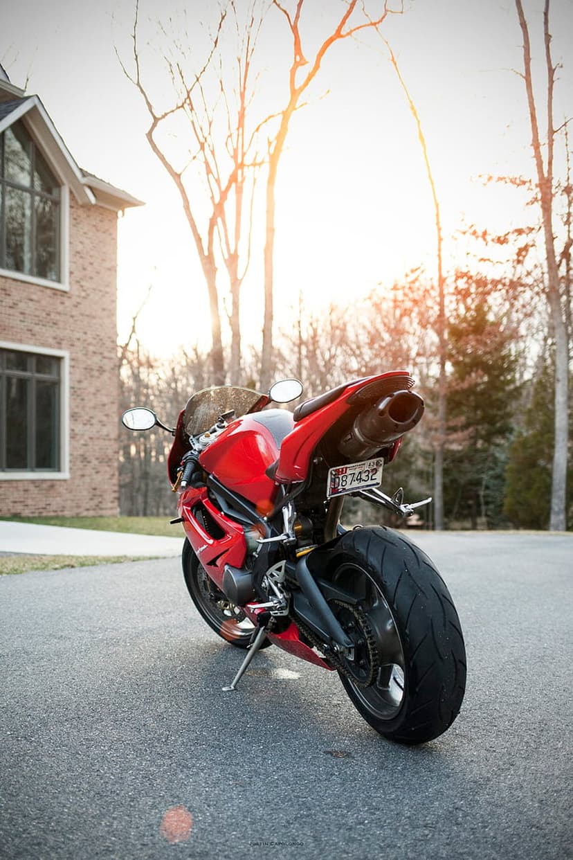 Red sport motorcycle parked on a driveway in front of a brick house with bare trees under a hazy, bright sky.