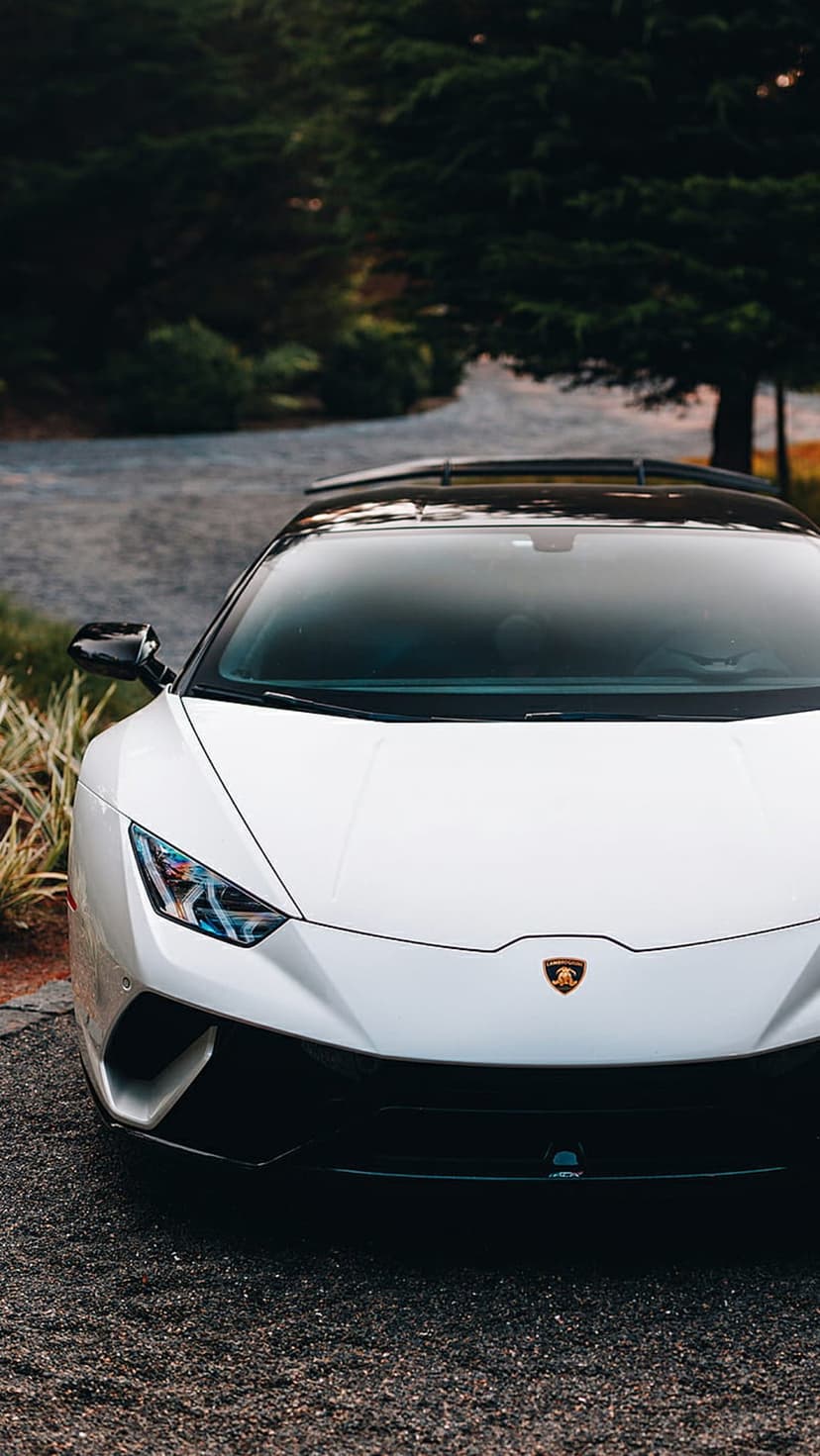 White Lamborghini Huracan on a dark gravel driveway with green foliage, framed for a vertical phone screen.