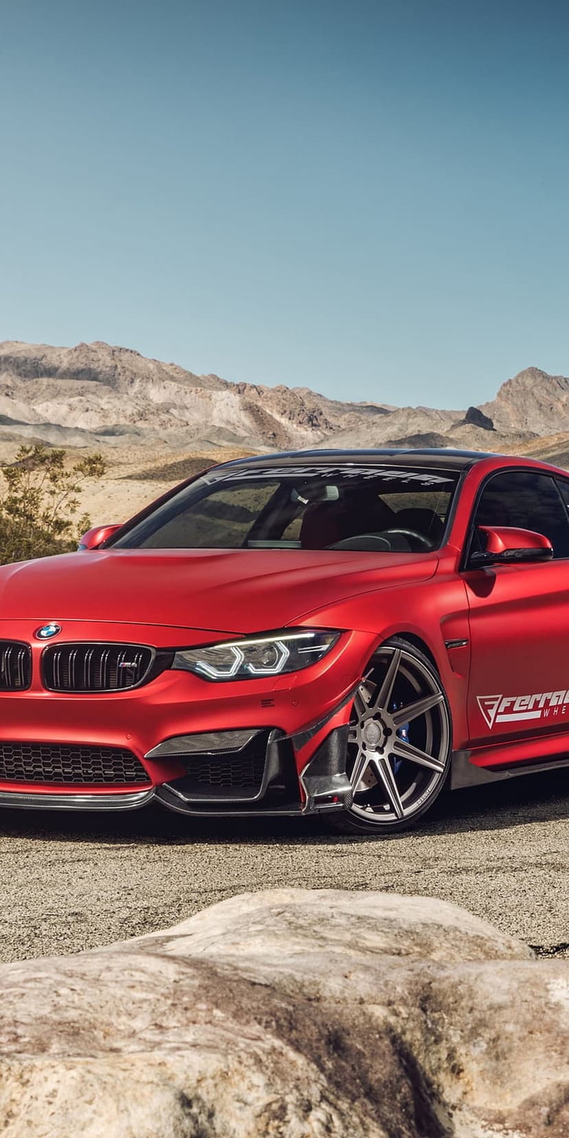 Red BMW M4 coupe parked on a gravel desert path with rocky mountains and a clear blue sky in the background.