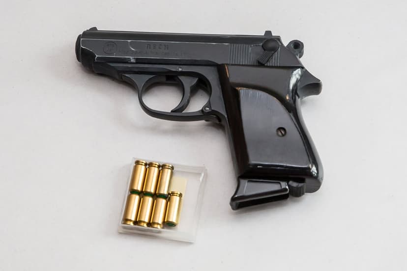 Walther PPK handgun beside a six-round pack of brass cartridges on a smooth, brightly lit white studio surface.