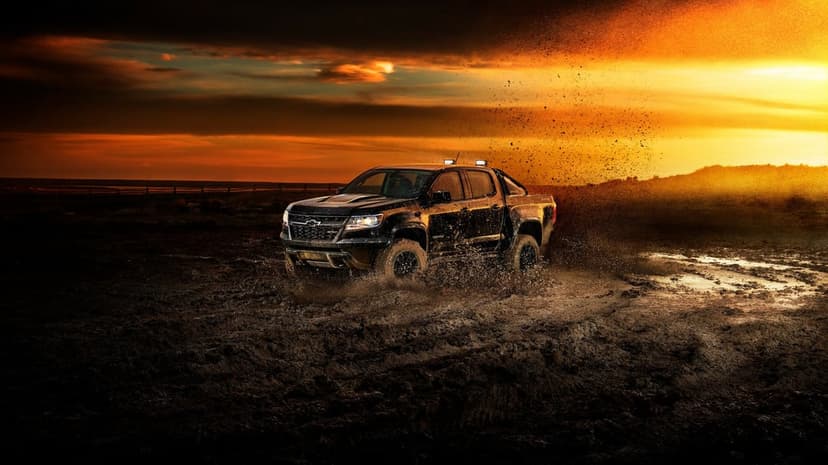 Black off-road truck splashing through a muddy field under a vibrant orange and red sunset sky.