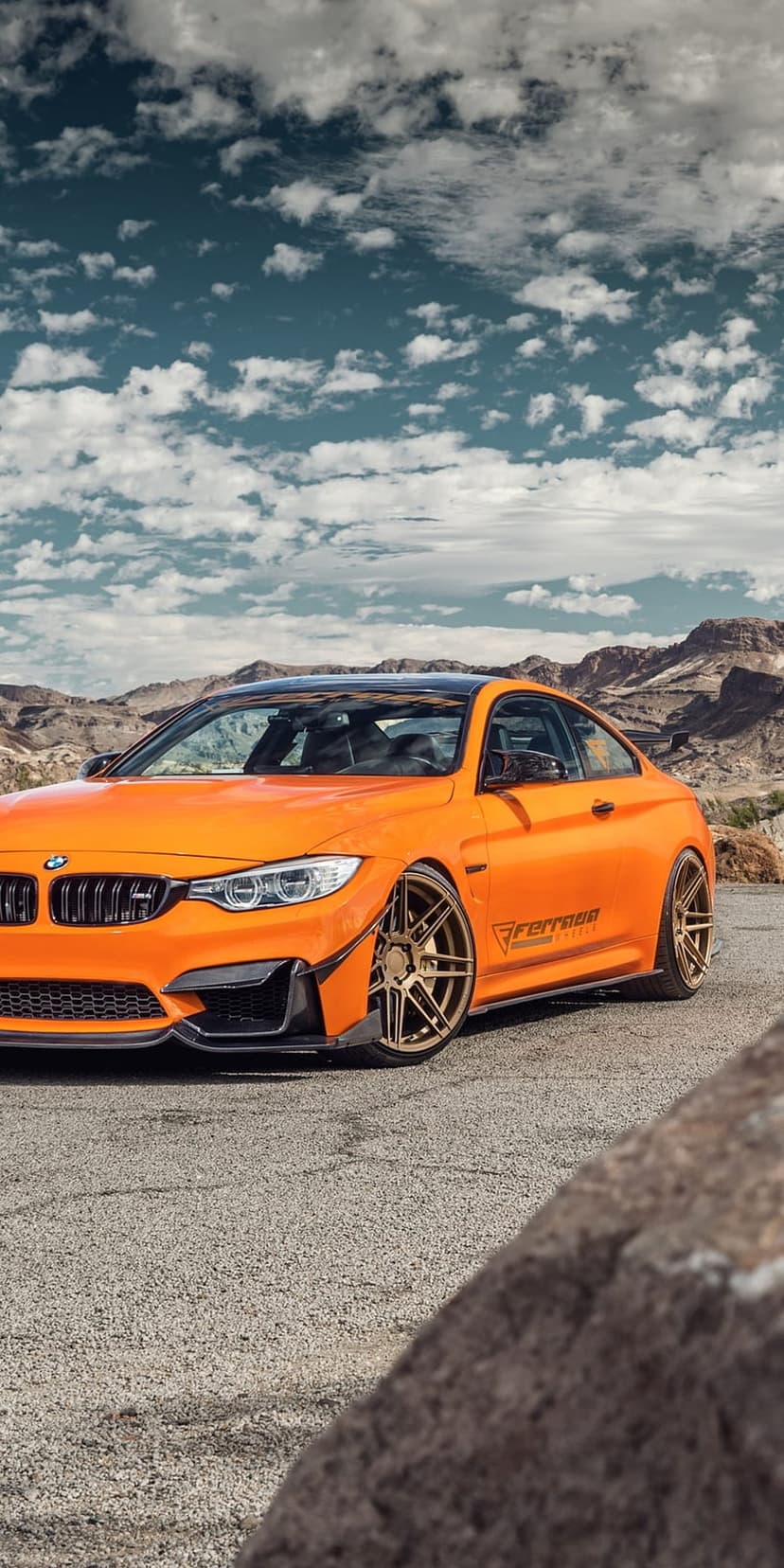 Orange BMW M4 coupe with bronze wheels parked on a desert road against a backdrop of mountains and cloudy skies.