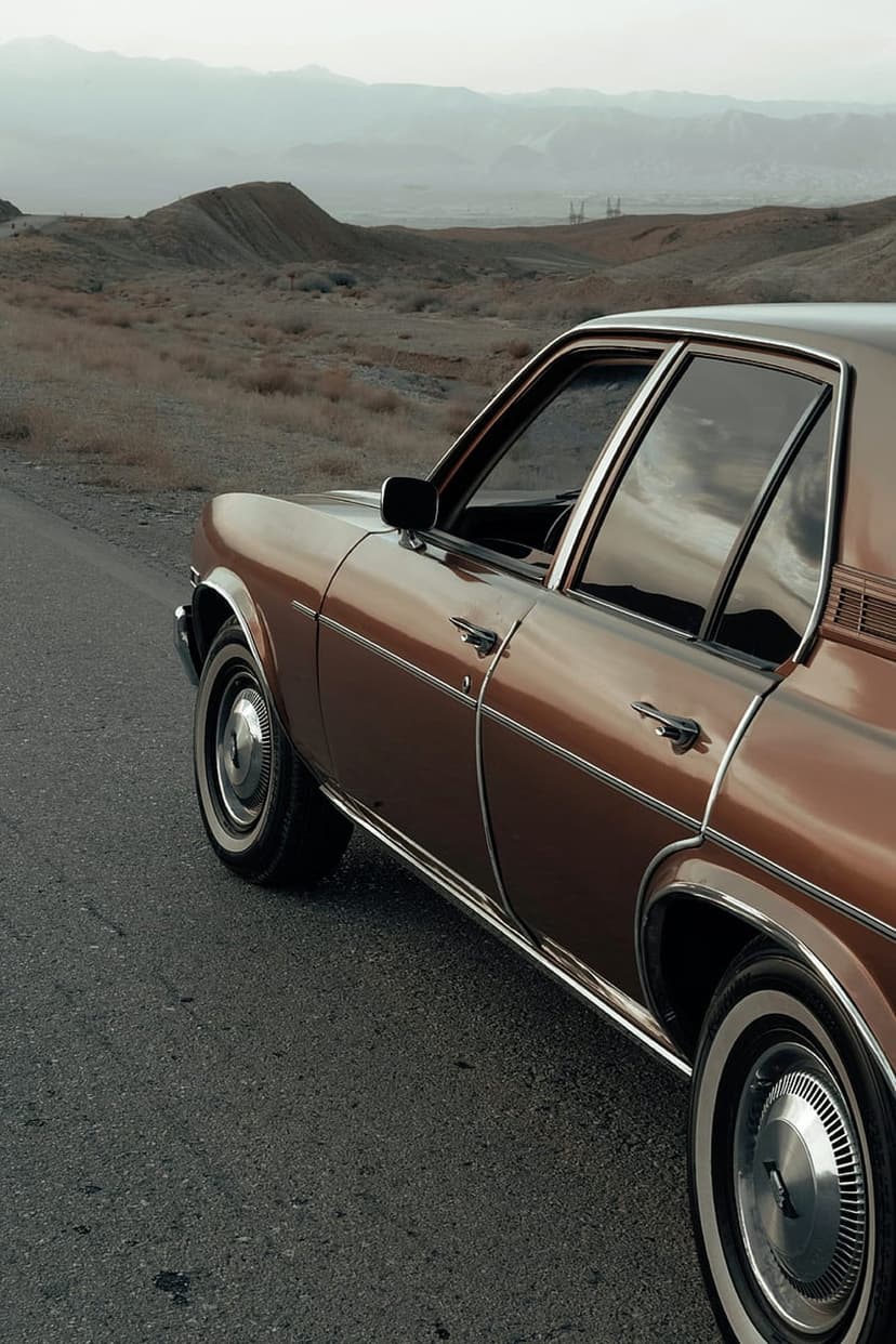 Vintage brown sedan with chrome details and whitewall tires parked on a desert highway under a cloudy sky.