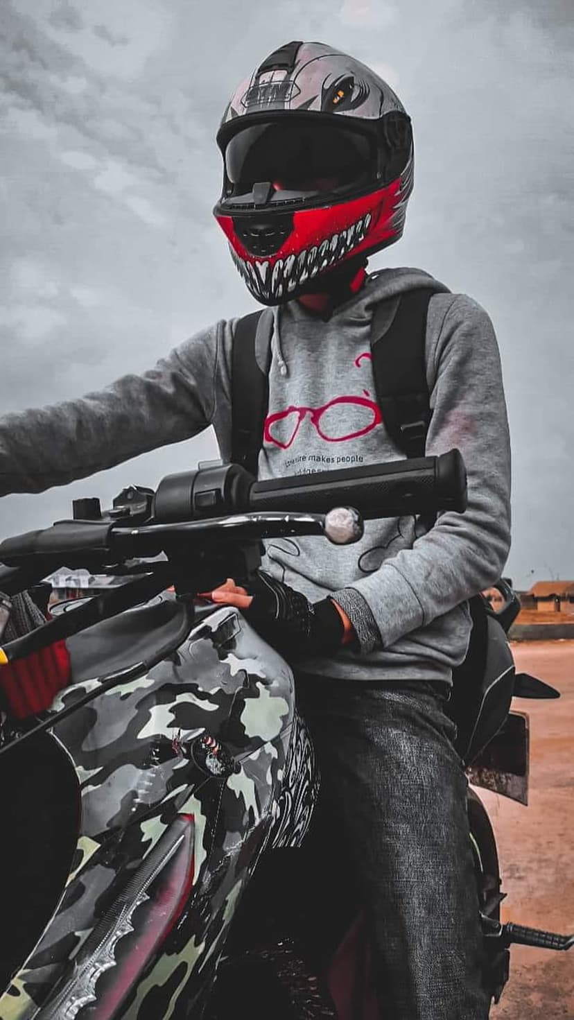 Biker in a shark-shaped helmet and grey hoodie sits on a camouflage motorcycle beneath a dramatic overcast sky.