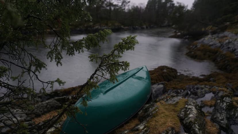 Green canoe rests on a mossy rocky shore beside a dark lake while rain falls in a misty, overcast pine forest.