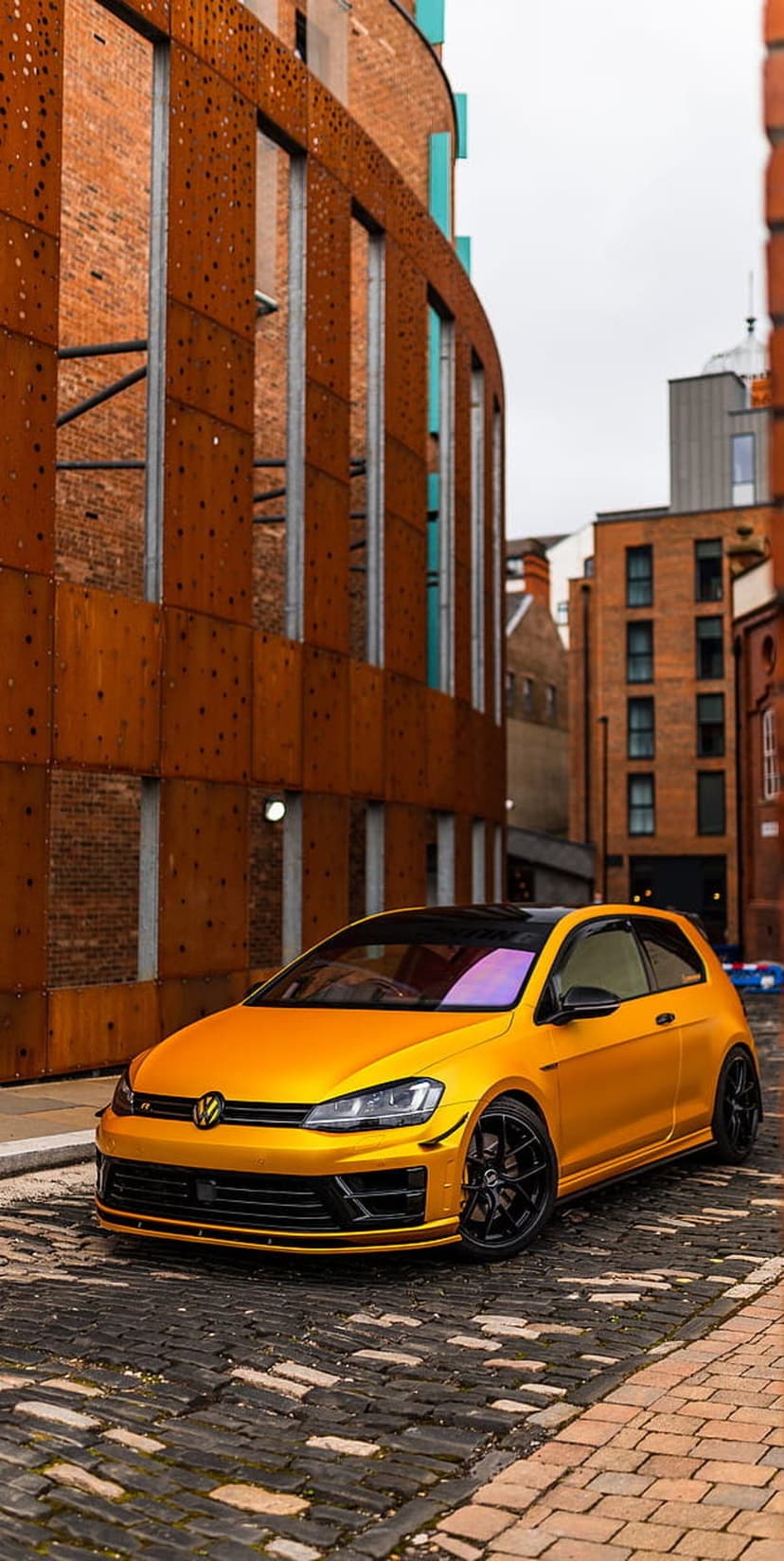 Yellow Volkswagen Golf GTI parked on textured cobblestones in front of industrial brick and metal buildings.