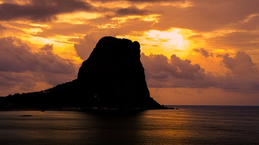 Volcanic island silhouette under a golden orange sunset with dramatic clouds reflected on dark ocean waves.