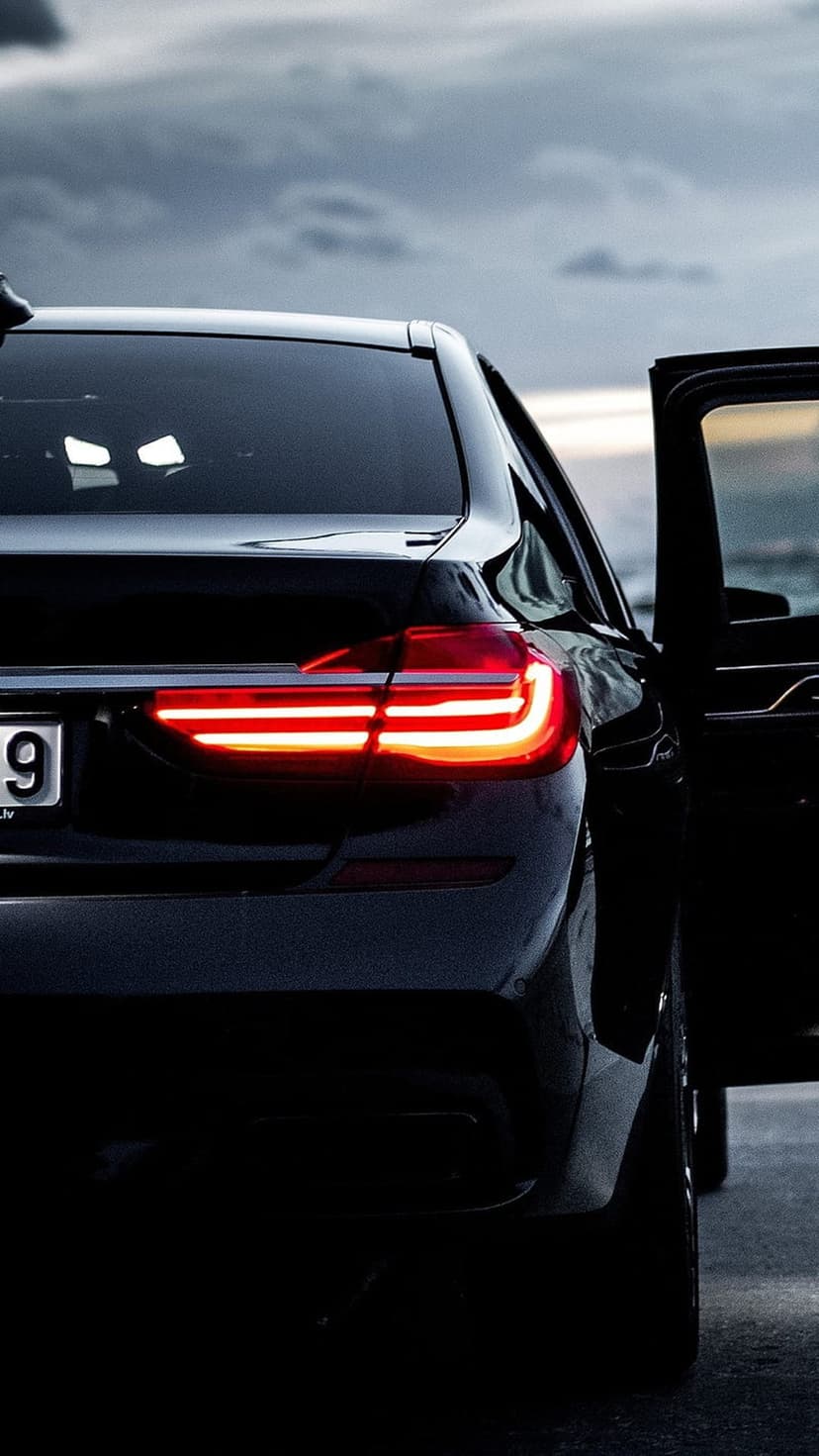 Black luxury sedan rear view with lit taillights and an open door parked near a coastal ocean horizon at dusk.