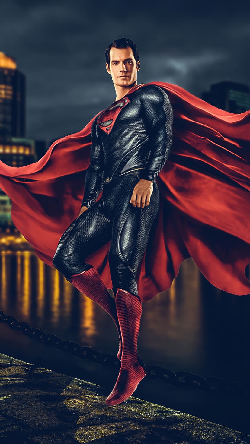 Superman stands on a rocky cliff overlooking a dark city with a long red cape billowing in the night wind.