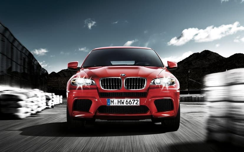 Red BMW X6 M SUV driving forward on a road with headlights on and motion blur against a blue sky.