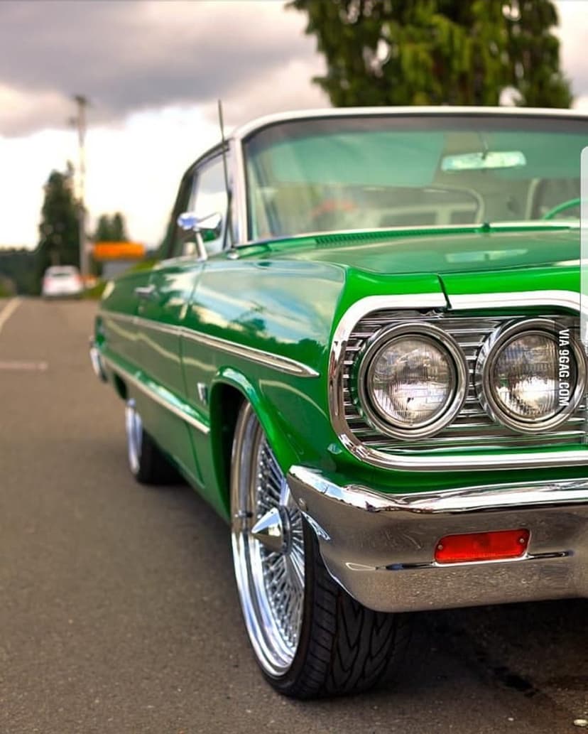 Green classic car with chrome grille and wire wheels parked on a road under a cloudy sky from a low angle.