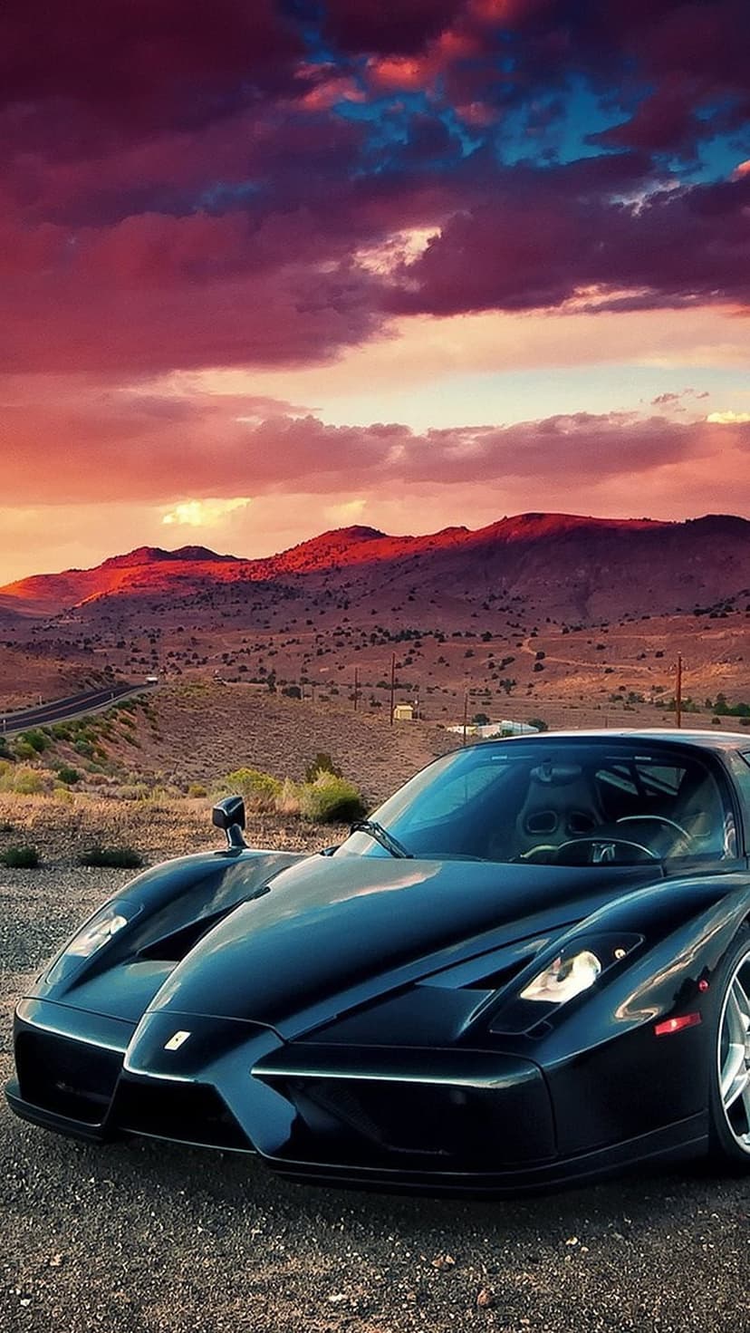 Black Ferrari Enzo parked on a desert highway under a vivid orange and purple sunset sky with distant mountains.