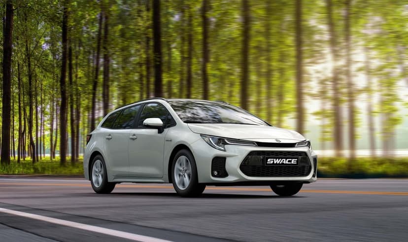 White Suzuki Swace hybrid wagon driving on a road with motion-blurred forest trees and warm sunlight.