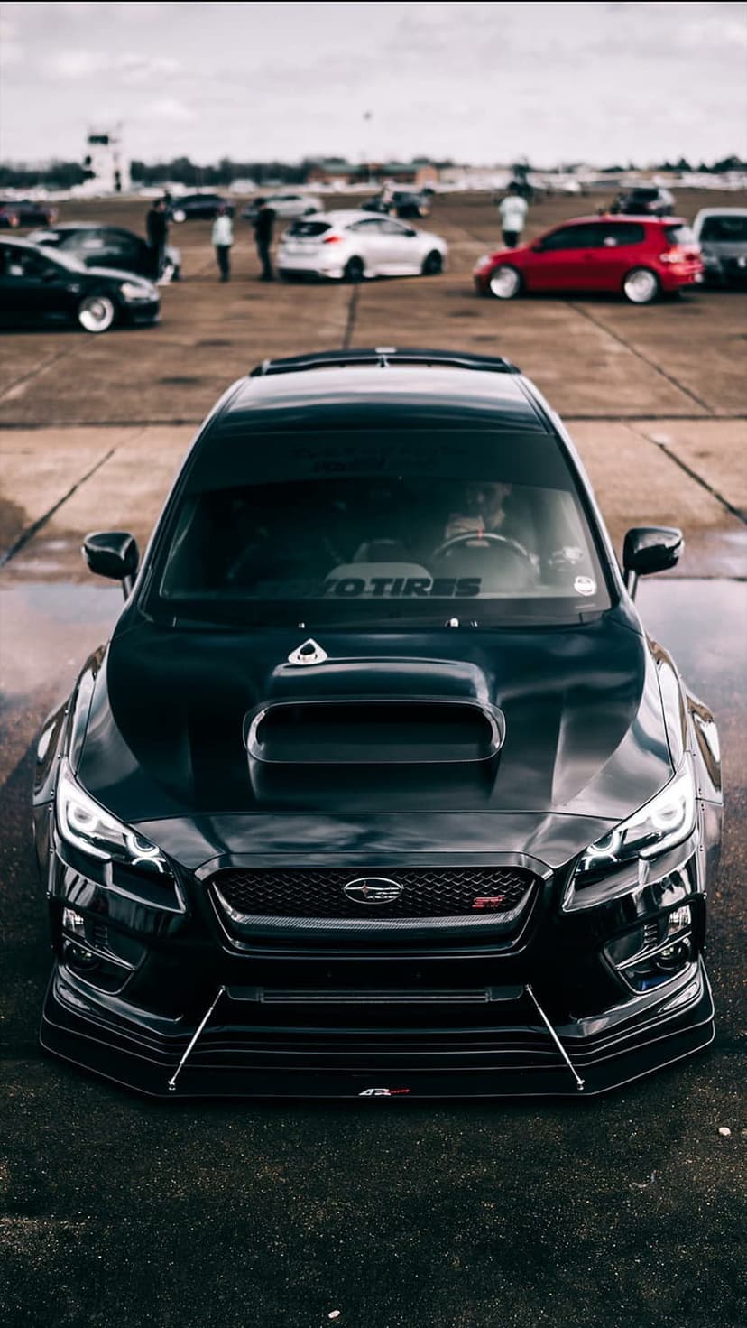 Black Subaru WRX STI car with a hood scoop parked on wet asphalt during an overcast automotive meet.