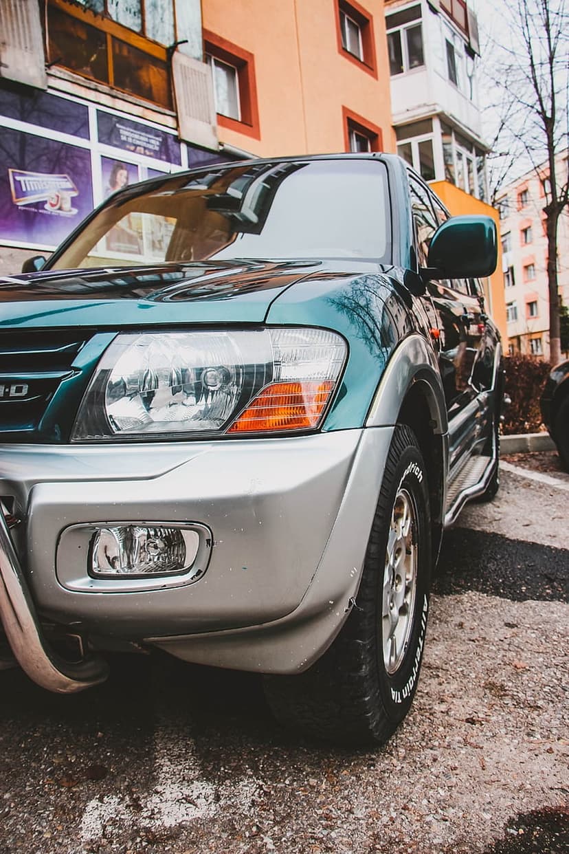 Dark green SUV wheel and headlight with all-terrain tires parked near orange apartment buildings in the city.
