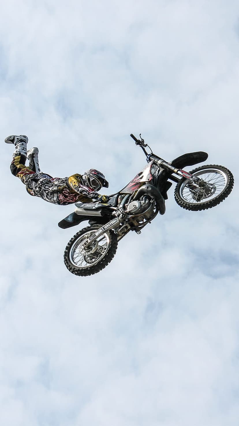 Motocross rider in mid-air performing a stunt on a dirt bike against a backdrop of soft white clouds in the sky.