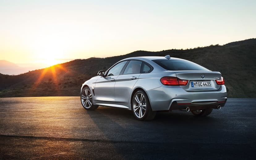 Rear view of a silver BMW 4 Series parked on a road at sunset with mountains and a sun flare background.