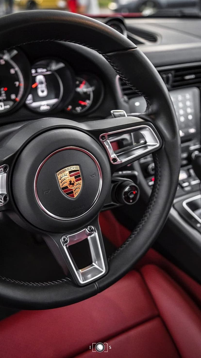Porsche steering wheel with gold crest and black leather in front of a vibrant red luxury car interior.