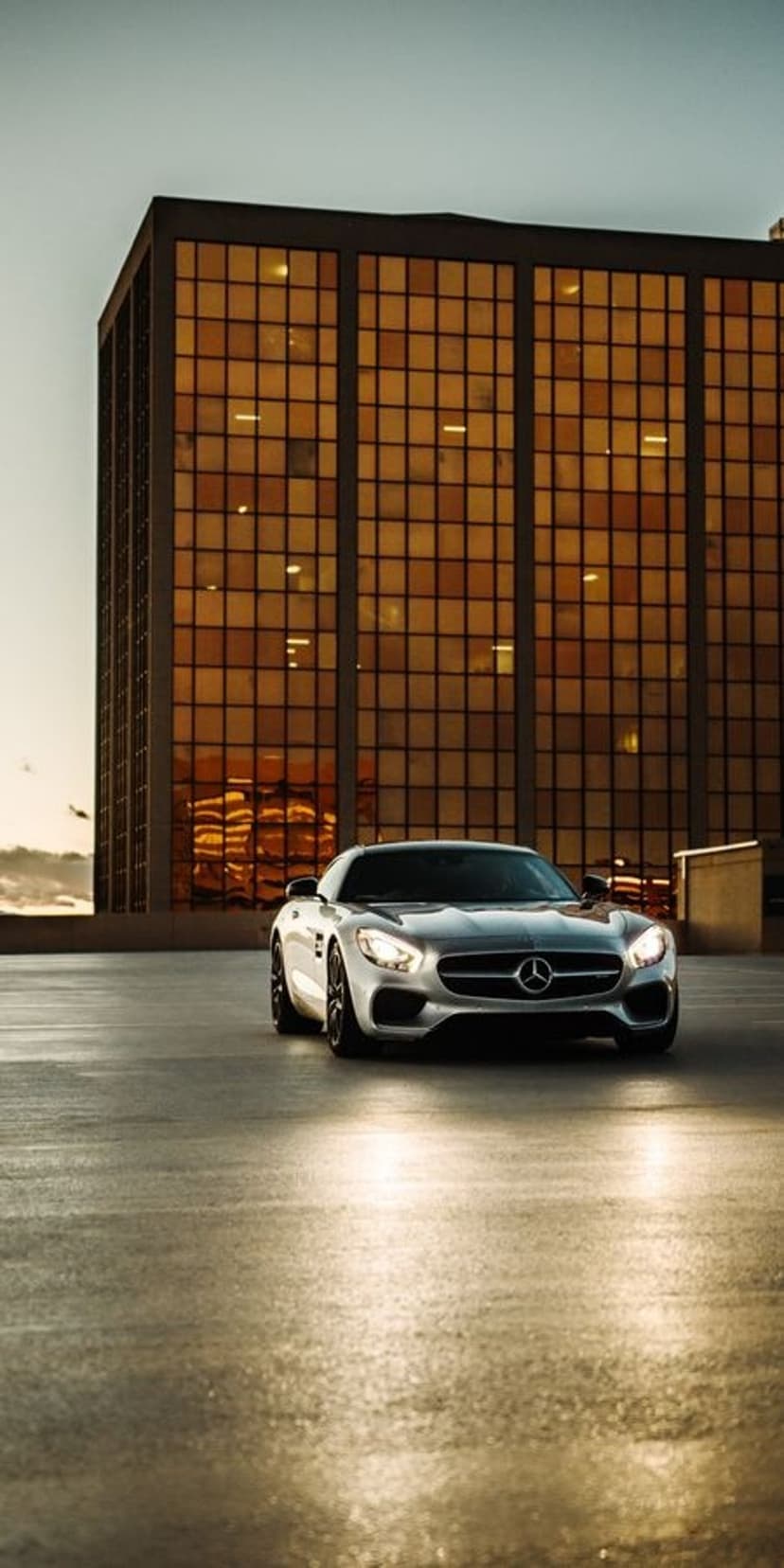 Silver Mercedes AMG GT parked on reflective concrete with headlights on before a golden glass building at sunset.
