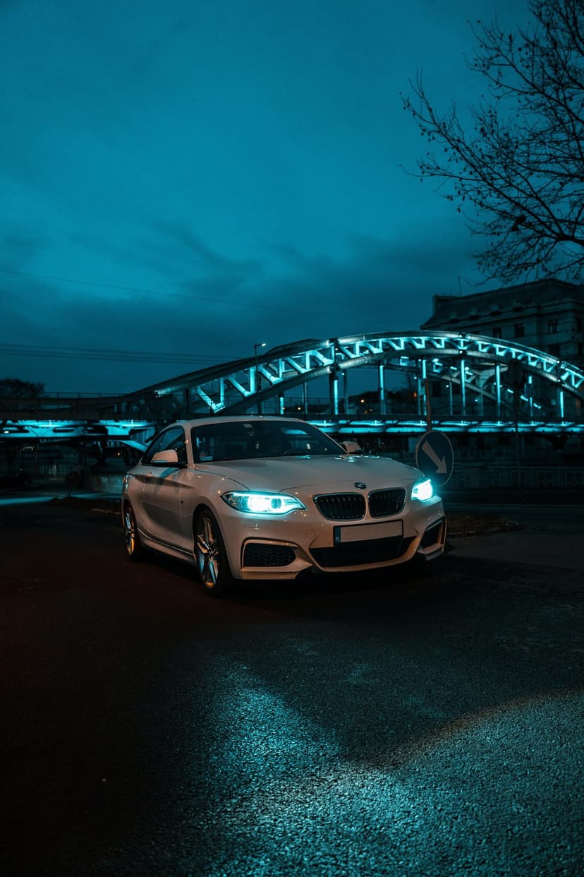 White BMW luxury sedan on a wet road at night with glowing blue headlights and a lit bridge in the background.