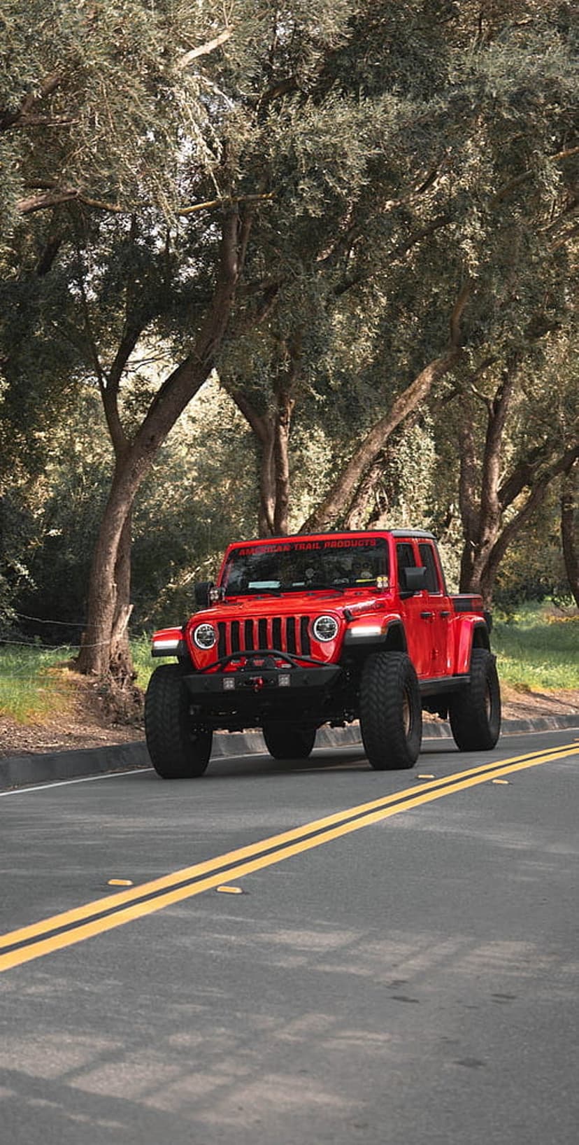 Red Jeep Gladiator pickup truck drives on a forest road with sunlight filtering through the leafy canopy.