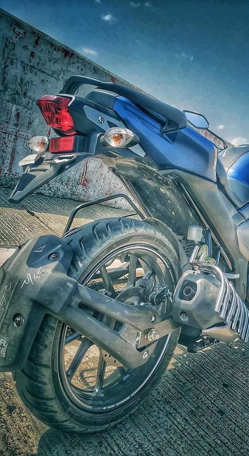 Blue motorcycle rear wheel with textured tire and red tail light against a concrete wall and cloudy blue sky.