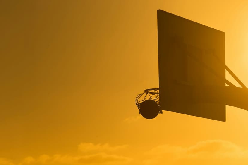 Basketball hoop and backboard silhouetted against a vibrant orange and yellow sunset sky during golden hour.