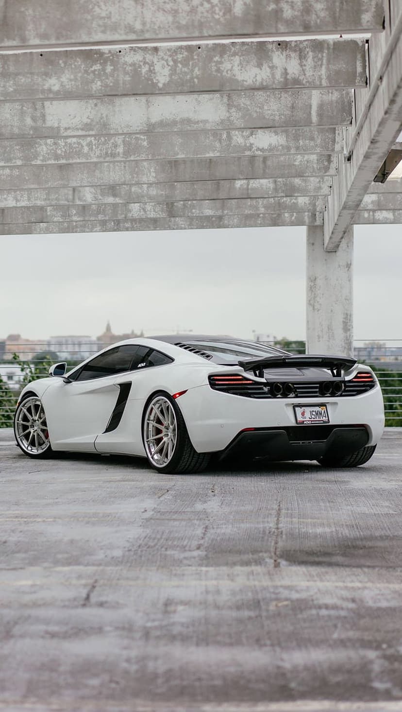 White McLaren supercar with a large rear wing parked on concrete under a weathered urban bridge structure.