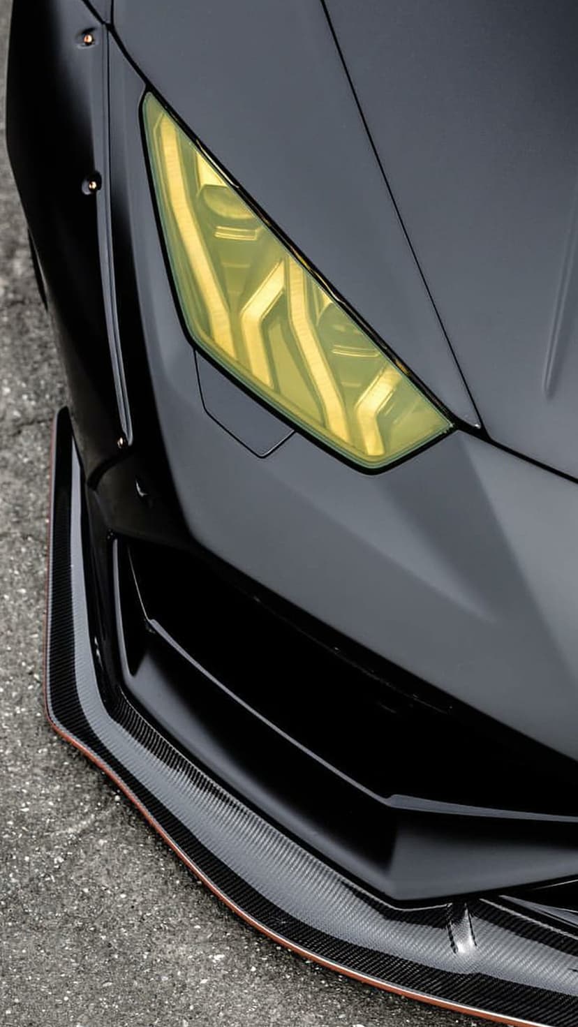 Lamborghini yellow LED headlight and carbon fiber bumper with copper trim on a matte black supercar body.