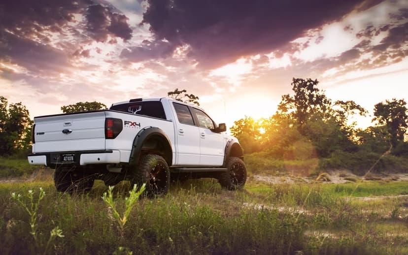 Lifted white pickup truck in a grassy field at sunset with dramatic purple clouds and golden lighting.