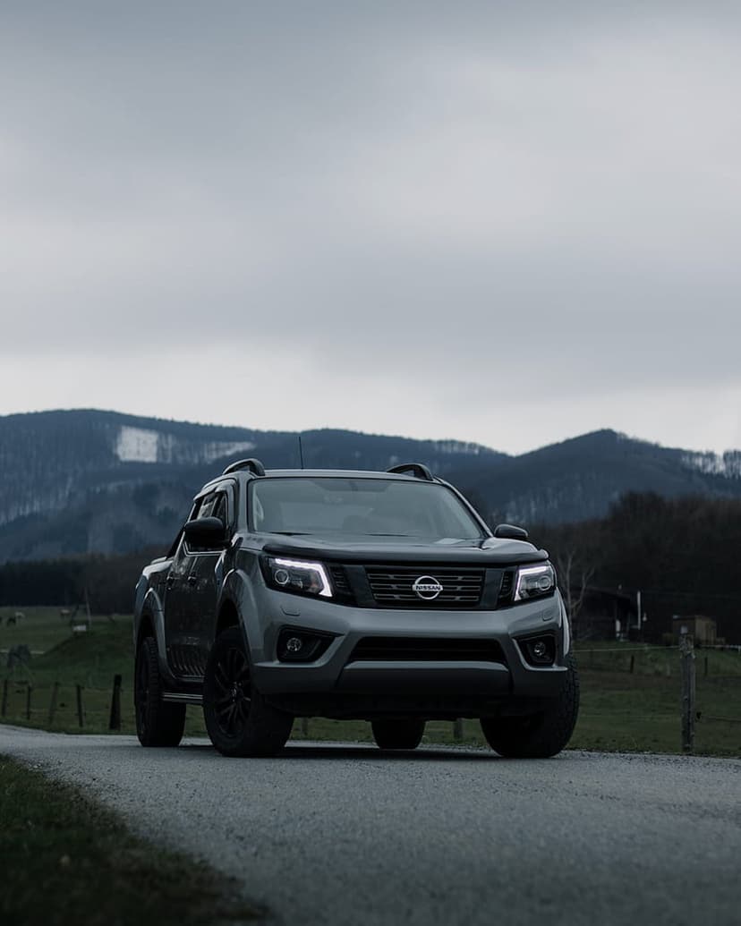 Nissan Navara pickup truck with glowing headlights on a gravel road against a cloudy, mountainous backdrop.