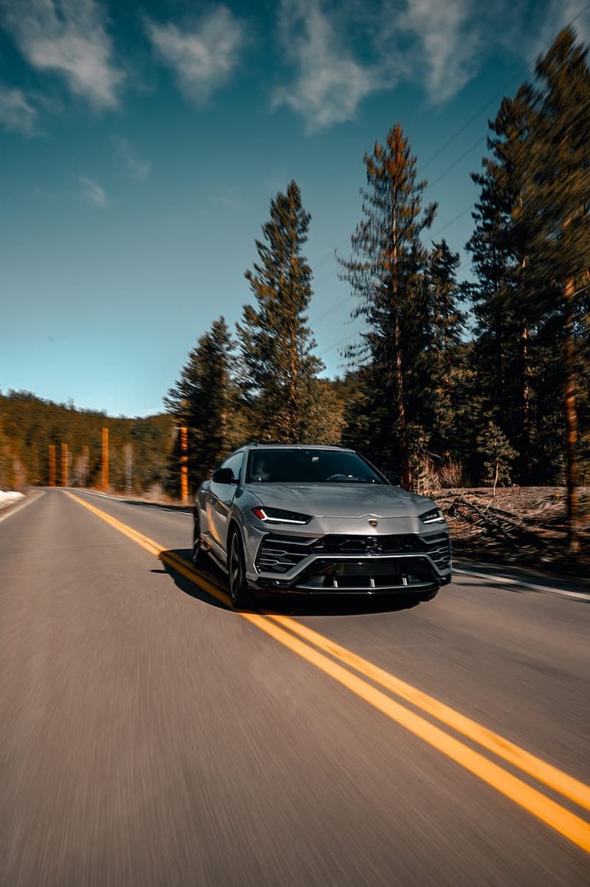 Silver Lamborghini Urus SUV speeds past evergreen trees on a mountain road with motion blur on the pavement.
