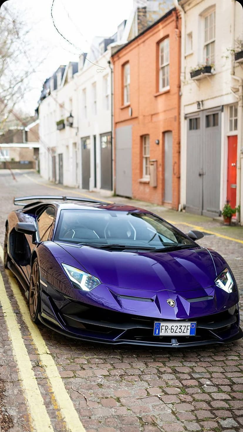 Purple Lamborghini Aventador with glowing LED headlights parked on a gray cobblestone street between old buildings.