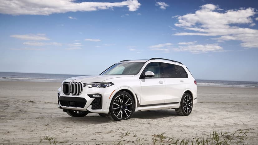 White BMW X7 luxury SUV parked on a sandy beach under a blue sky with soft white clouds and ocean views.