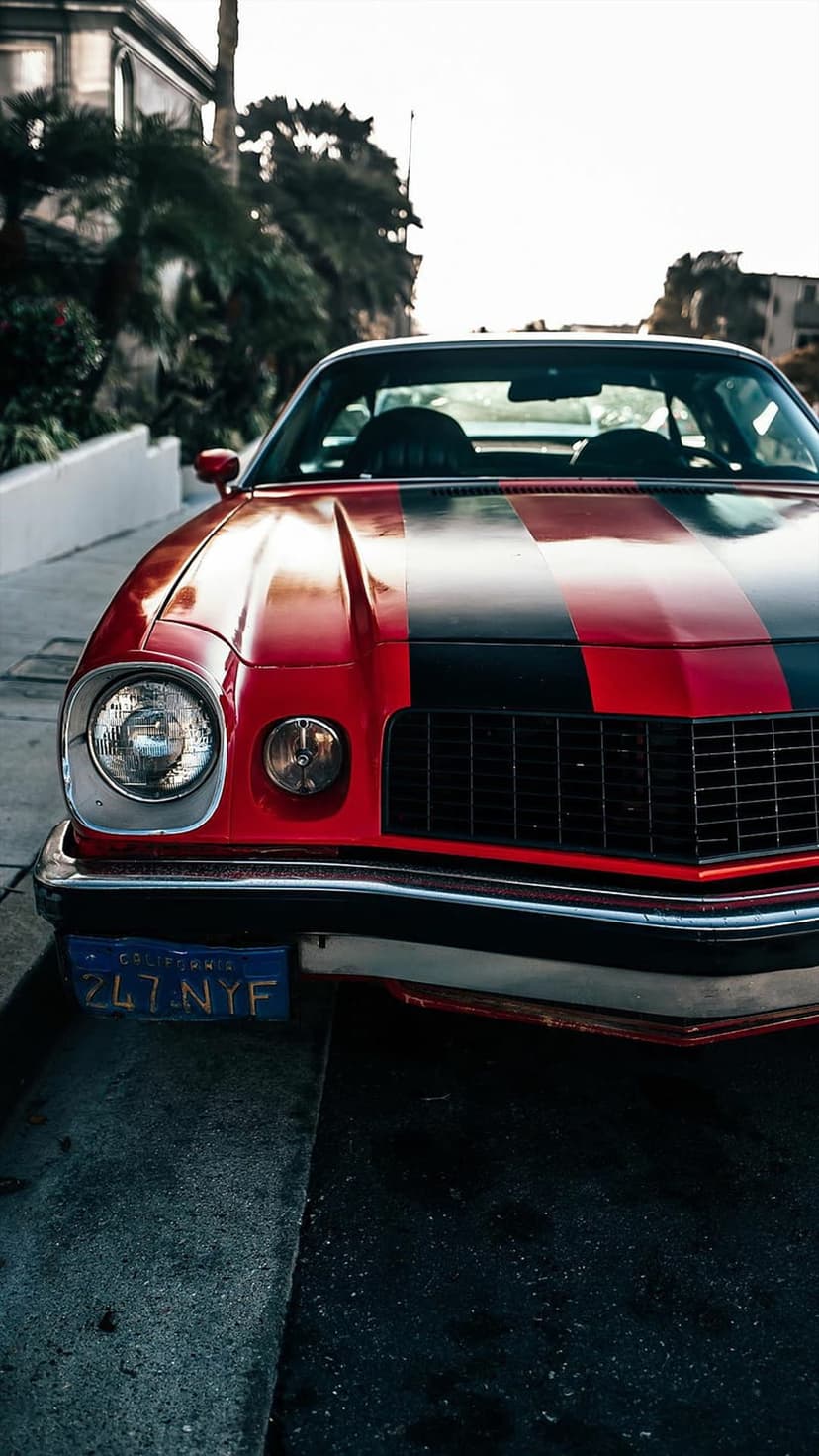Red vintage Camaro with black stripes parked on a sunlit street with palm trees and a bright sky backdrop.