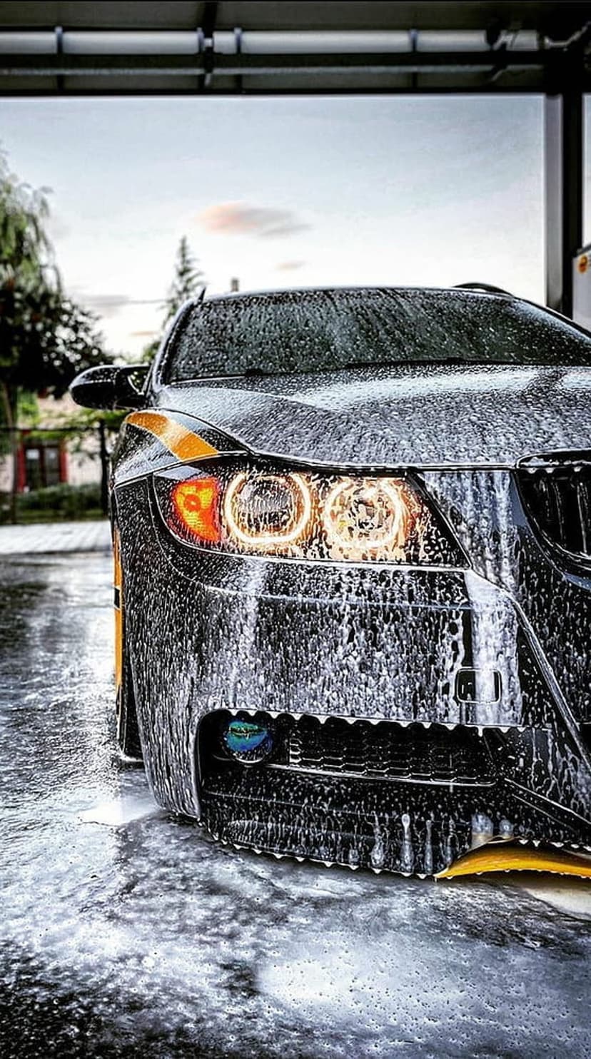 Black luxury car front end covered in white soap suds featuring glowing headlights and bright yellow accents.