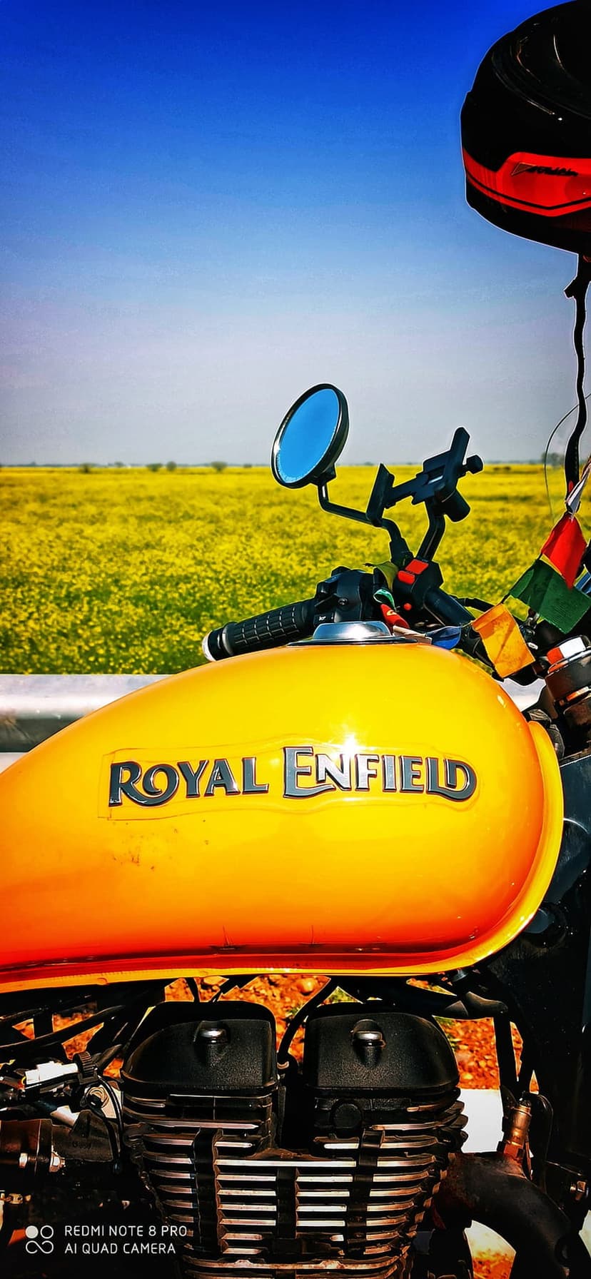 Orange Royal Enfield motorcycle parked beside a vast field of yellow flowers under a clear blue summer sky.