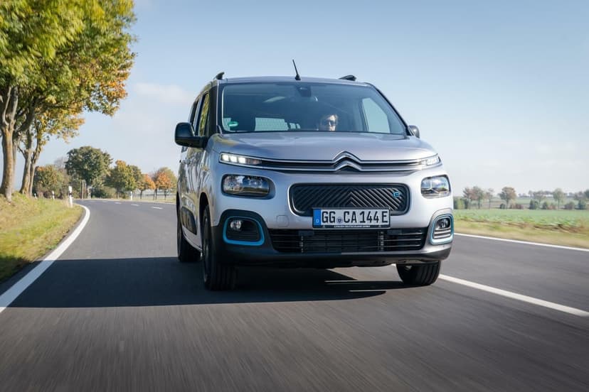 Silver Citroën electric van driving on a paved road through green fields under a clear blue sky with motion blur.