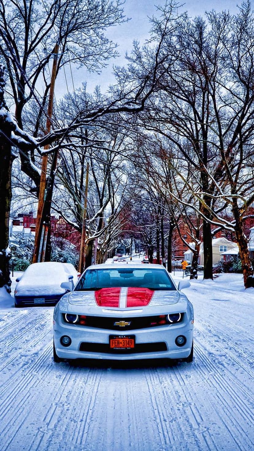 White Camaro with red racing stripes parked on a snowy street under bare trees with glowing headlights visible.