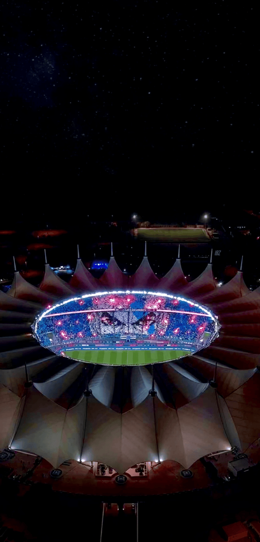 Football stadium with a glowing tent-roof and green field shines under a starry night sky from an aerial view.