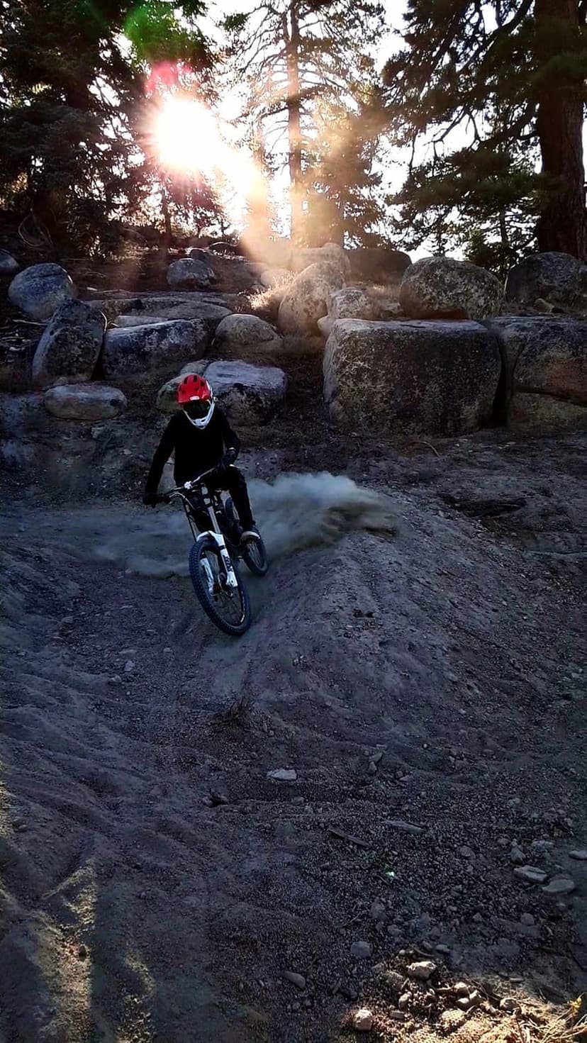 Mountain biker in a red helmet kicks up a dust cloud while riding downhill through a sunlit, rocky forest trail.