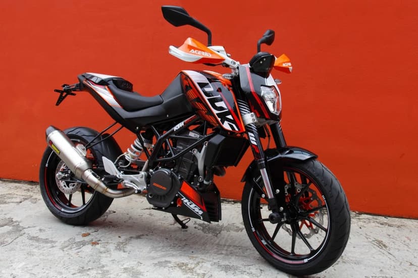 KTM Duke motorcycle with Akrapovic exhaust parked against a solid vibrant orange wall on a concrete surface.
