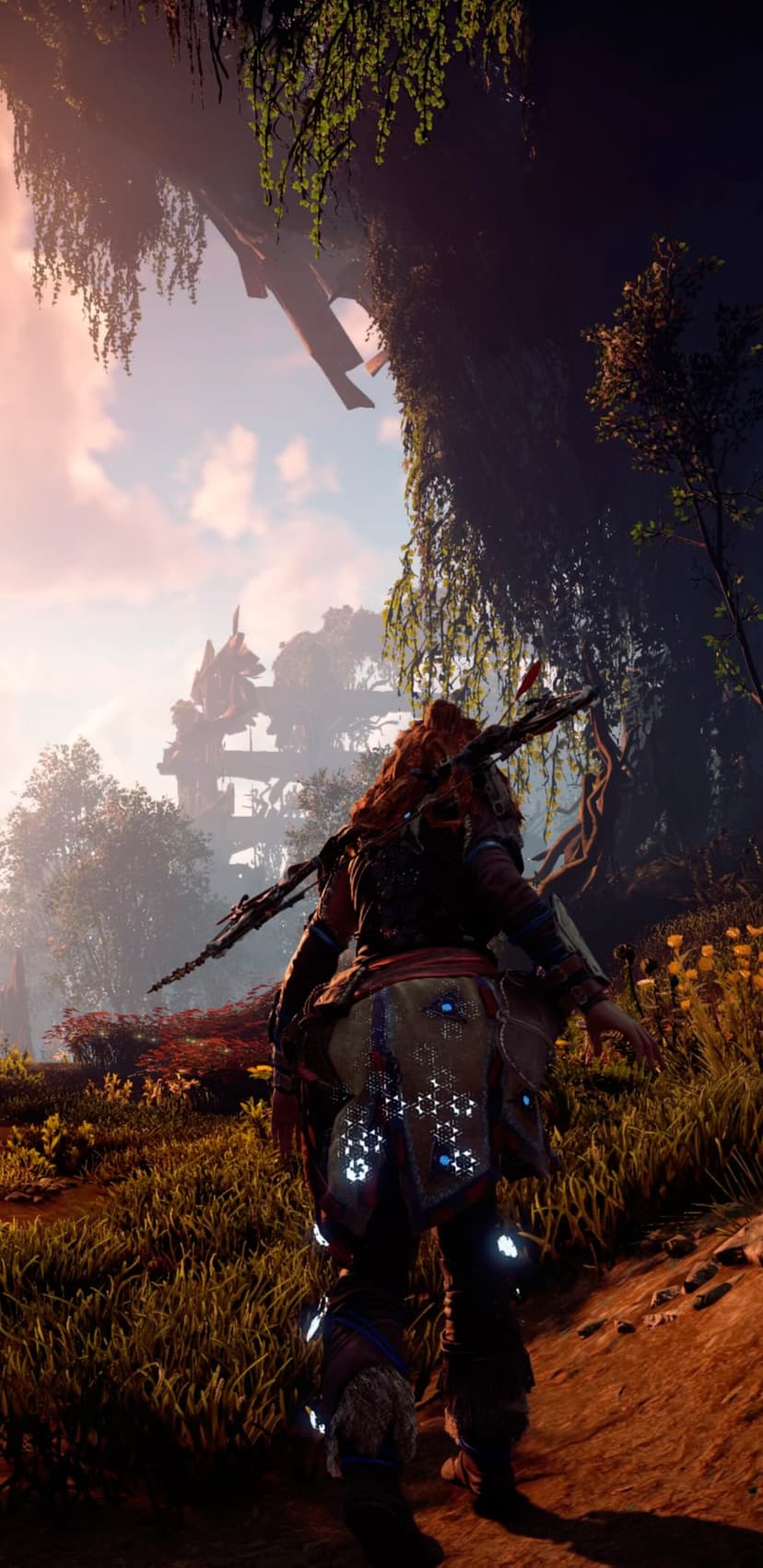 Aloy with red hair and glowing blue armor walks through a sunlit forest toward distant mechanical ruins.
